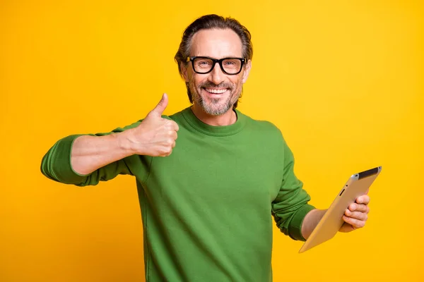 Foto de hombre alegre mantenga la tableta levantar el pulgar hacia arriba sonrisa blanca usar gafas jersey verde aislado color amarillo fondo — Foto de Stock