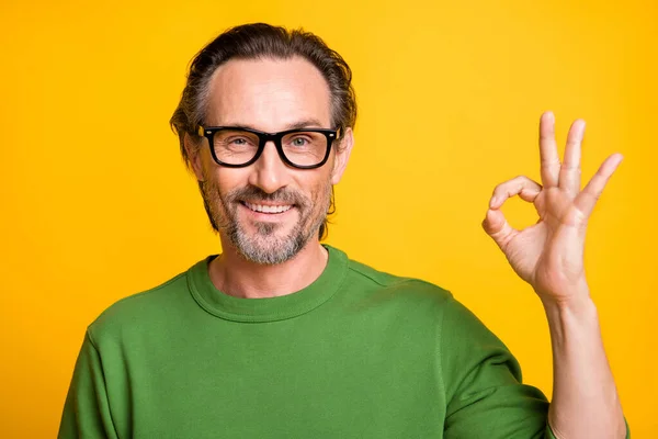 Fénykép vidám szakállas srác ujjak mutatják okey viselet szemüveg zöld pulóver izolált sárga színű háttér — Stock Fotó