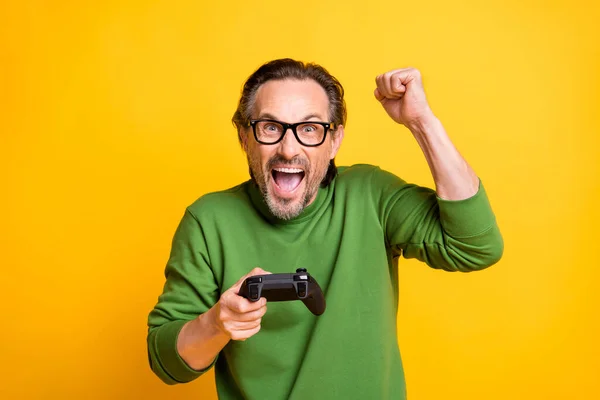 Foto de chico triunfante alegre mantenga gamepad ganar juego levantar puño usar gafas suéter verde aislado color amarillo fondo — Foto de Stock