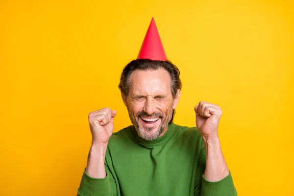 Foto de homem barbudo animado levantar punhos fechar os olhos esperar desgaste cone chapéu verde pulôver isolado cor amarela fundo — Fotografia de Stock