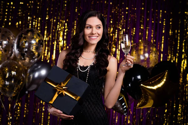 Foto de adorable linda mujer joven vestida traje negro sosteniendo regalo beber vino buscando espacio vacío aislado oro brillo fondo —  Fotos de Stock
