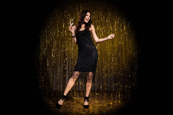 Full size photo of brown optimistic lady stand with glass look empty space wear black dress isolated on dark gold color background in stage light — Stock Photo, Image