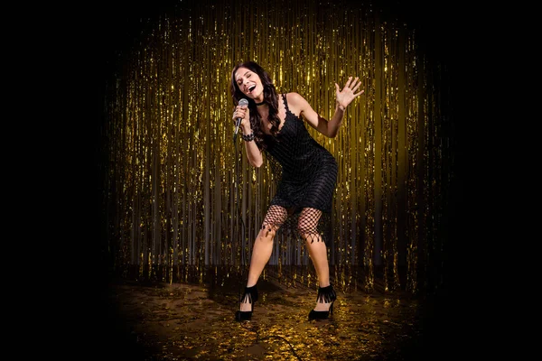 Foto in voller Größe von braunen optimistischen Dame stehen singen in Mikrofon tragen schwarze Kleid Schuhe isoliert auf dunkelgoldenen Farbhintergrund in Bühnenlicht — Stockfoto