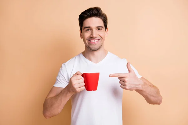 Портрет оптимістичної брюнетки чоловік вказує на чашку носити білу футболку ізольовано на бежевому кольоровому фоні — стокове фото