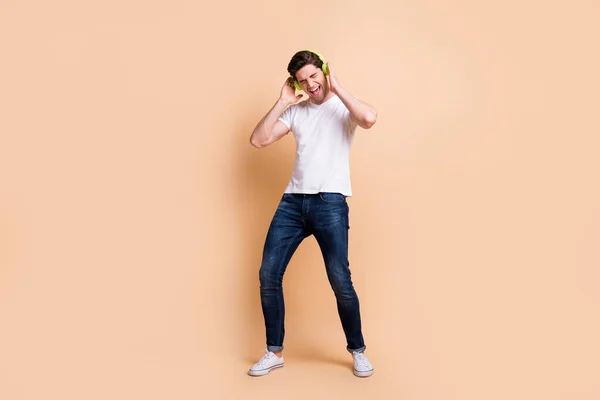 Plná velikost fotografie optimistický brunet muž poslouchat hudbu nosit bílé tričko džíny tenisky izolované na béžové barvy pozadí — Stock fotografie