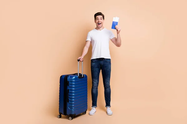 Фотографія враженого брюнетки чоловік тримає сумку з квитками, одягнені в футболки джинси кросівки ізольовані на бежевому фоні — стокове фото