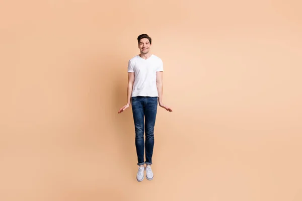 Full size φωτογραφία του αισιόδοξου brunet άνθρωπος άλμα φορούν t-shirt τζιν sneakers απομονώνονται σε μπεζ φόντο — Φωτογραφία Αρχείου