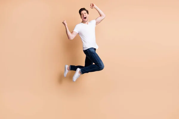 Полноразмерная фотография профиля ура-брюнета, прыгающего в джинсах и футболках на бежевом фоне — стоковое фото