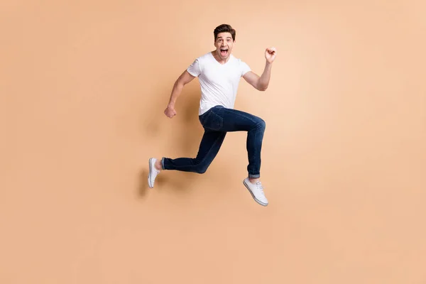 Teljes méretű profil fotó hurrá brunet férfi ugrás futás kiabálni viselni póló farmer tornacipő elszigetelt bézs háttér — Stock Fotó