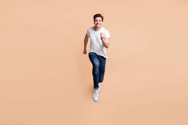Foto de tamaño completo de hombre morena optimista correr usar zapatillas vaqueros camiseta aislado en fondo de color beige —  Fotos de Stock