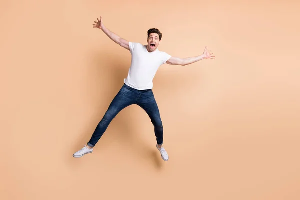 Full size photo of optimistic brunet man jump yell wear t-shirt jeans sneakers isolated on beige color background — Stock Photo, Image