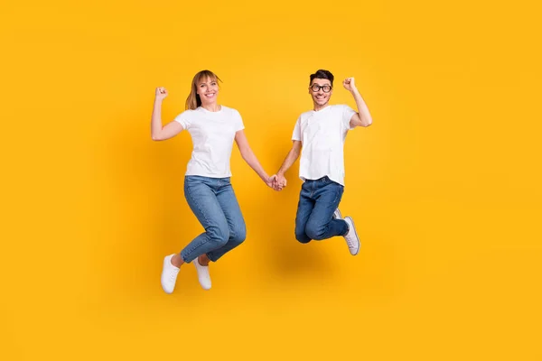 Volledige lengte foto van schattige zoete personen gekleed wit t-shirt springen hand in hand stijgende vuisten geïsoleerde gele kleur achtergrond — Stockfoto