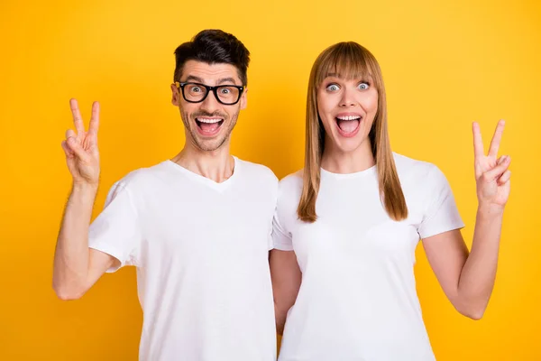 Fénykép aranyos lenyűgözött fiatal pár viselni fehér póló szemüveg mutatja v-jelek nyitott száj izolált sárga háttér — Stock Fotó