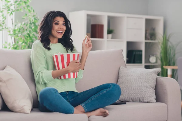 Portræt af attraktiv munter pige sidder på divan spise popcorn ser film komedie have det sjovt i flad lejlighed indendørs - Stock-foto