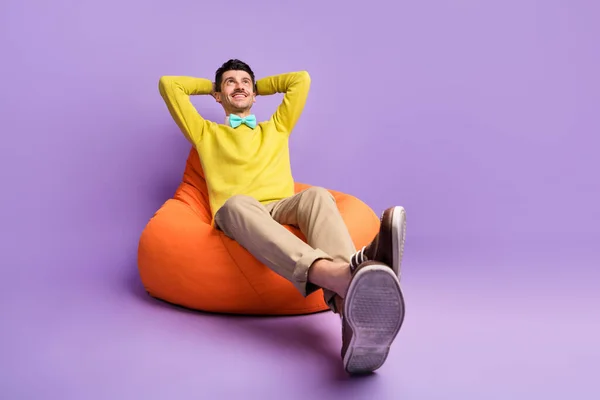 Full size foto di brunet ottimistico ragazzo sedersi su borsa mani sotto la testa usura giallo maglione pantaloni scarpe da ginnastica isolato su sfondo lilla — Foto Stock