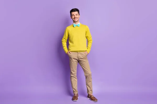 Foto de tamaño completo de tipo optimista morena de pie de las manos en los bolsillos usan suéter amarillo pantalones zapatillas aisladas sobre fondo lila —  Fotos de Stock