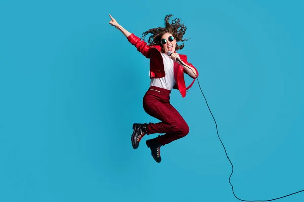 Full lengde portrett av en gal ung jente som hopper mens hun synger direkte tomrom åpen munn isolert på blå bakgrunn – stockfoto