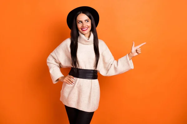 Фото милой очаровательной юной леди, одетой в бежевый пуловер, шляпа улыбается указывая пальцем пустое пространство изолированный оранжевый цвет фона — стоковое фото
