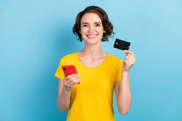 Bild av charmig flicka hålla mobiltelefon betalkort toothy leende bära gul t-shirt isolerad blå färg bakgrund — Stockfoto