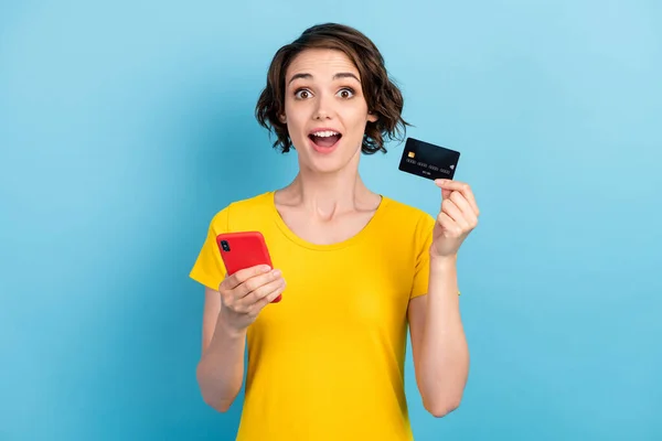 Fénykép a szép csodálkozó hölgy tartsa telefon hitelkártya nyitott száj kopás sárga póló elszigetelt kék háttér — Stock Fotó