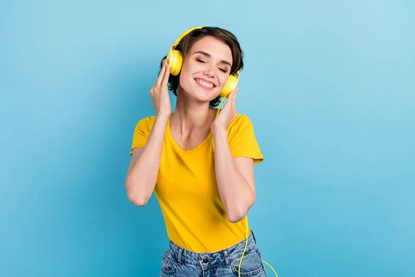Foto von charmanten jungen Mädchen mit geschlossenen Augen hören Musik tragen Kopfhörer gelbes T-Shirt isoliert blauer Hintergrund — Stockfoto
