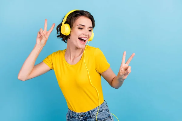 Foto de señora joven tienen espectáculo divertido v-signo escuchar música usar auriculares camiseta amarilla aislado color azul fondo — Foto de Stock