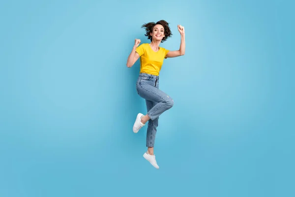 Foto de comprimento total de funky menina salto levantar punhos desgaste amarelo t-shirt jeans tênis isolado azul cor fundo — Fotografia de Stock