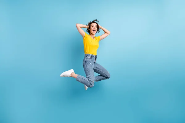 Foto de longitud completa de la señora divertida salto brazos cabeza boca abierta desgaste amarillo camiseta jeans zapatillas aislado color azul fondo —  Fotos de Stock