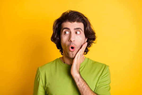 Foto de divertido chico sorprendido mejilla de la mano mirada lado espacio vacío desgaste verde camiseta aislado color amarillo fondo — Foto de Stock