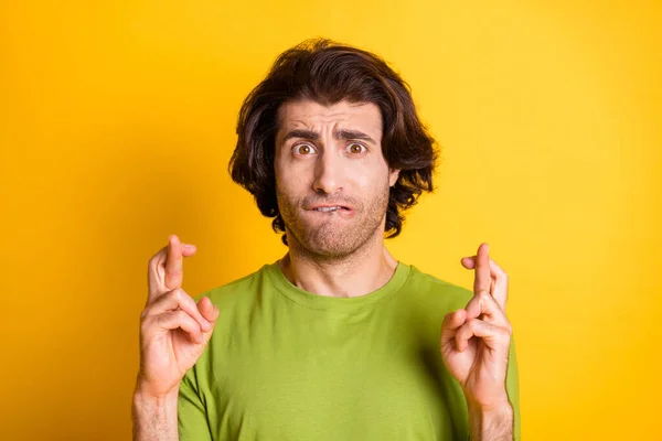 Foto de un joven preocupado morder el labio mostrando los dedos cruzados usar camiseta verde aislado color amarillo fondo — Foto de Stock