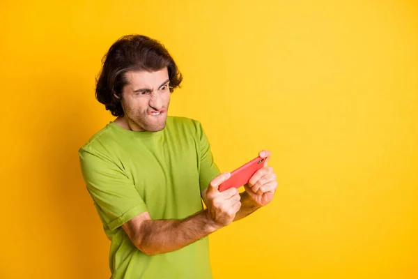 Foto van gek verslaafde man houden telefoon spelen spel bijten lip dragen groen t-shirt geïsoleerde gele kleur achtergrond — Stockfoto