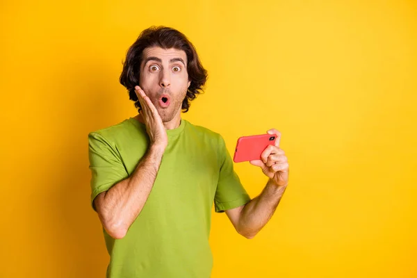 Foto van grappige geschokte man houden smartphone hand jukbeen dragen groen t-shirt geïsoleerde gele kleur achtergrond — Stockfoto