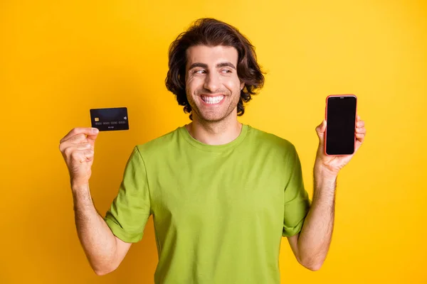 Foto van vrolijke man houd telefoon kijken scherm lege ruimte credit card dragen groen t-shirt geïsoleerde gele kleur achtergrond — Stockfoto