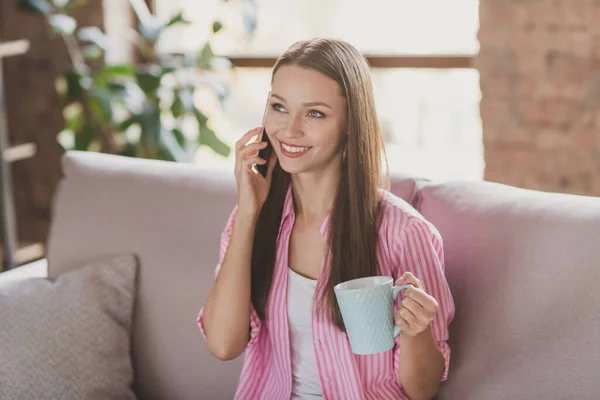 Fénykép nagyon édes fiatal nő öltözött csíkos ing ül kanapén beszél telefon ivás tea beltéri szobában — Stock Fotó