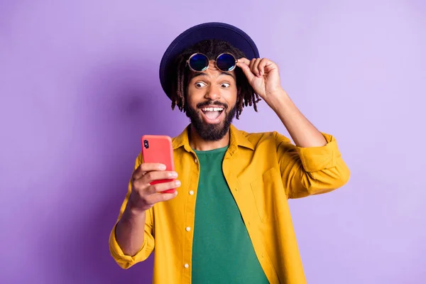 Foto di impressionato divertente pelle scura ragazzo mano tocco sunglass sguardo telefono come abbonarsi isolato su sfondo di colore viola — Foto Stock