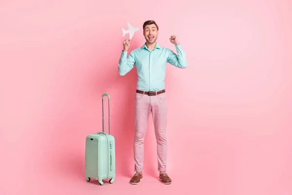 Full length body size photo of young man with suitcase keeping airplane figure gesturing like winner isolated on pastel pink color background — Zdjęcie stockowe