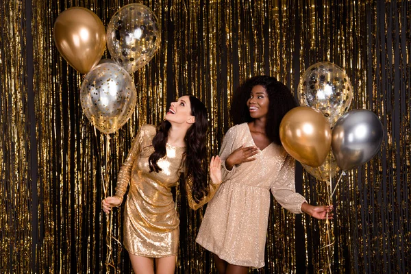 Foto de dos optimistas morena rizado peinado damas con globos se ven espacio vacío desgaste vestido aislado sobre fondo de color oro —  Fotos de Stock