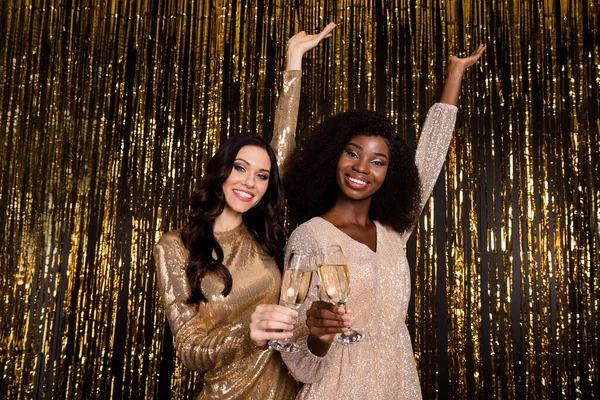 Photo of two lovely girlfriends hold glasses sparkling wine raise hands wear glossy outfit isolated tinsel background — Stock Photo, Image