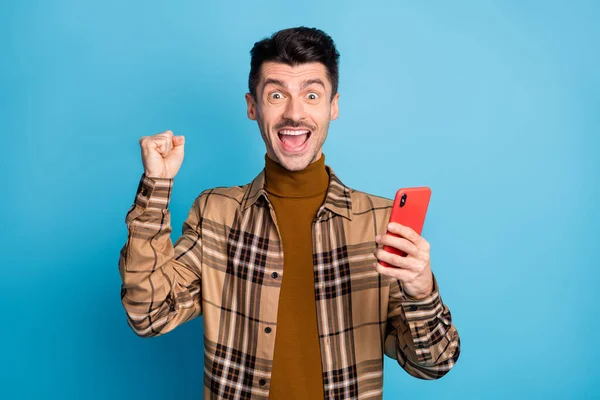 Foto di giovane felice eccitato pazzo positivo allegro uomo tenere il telefono alzare il pugno in vittoria isolato su sfondo di colore blu — Foto Stock