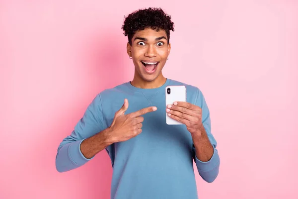 Foto de chico morena agradable impresionado punto teléfono desgaste suéter azul aislado sobre fondo de color rosa pastel — Foto de Stock