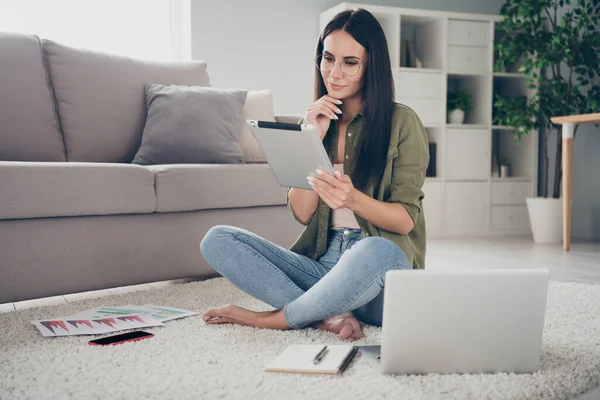 Full size photo of attractive positive lady sit on floor barefoot hand on chin look minded tablet working home indoors — Stock fotografie