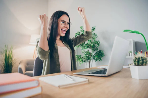 Фото молодой счастливой позитивной взволнованной сумасшедшей улыбающейся веселой женщины в очках, работающей в ноутбуке, поднимающей кулаки в победу дома — стоковое фото