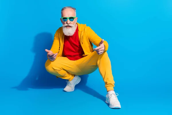 Retrato de comprimento total da pessoa idosa satisfeita agachamento braços dedos mostrando isolado no fundo de cor azul — Fotografia de Stock