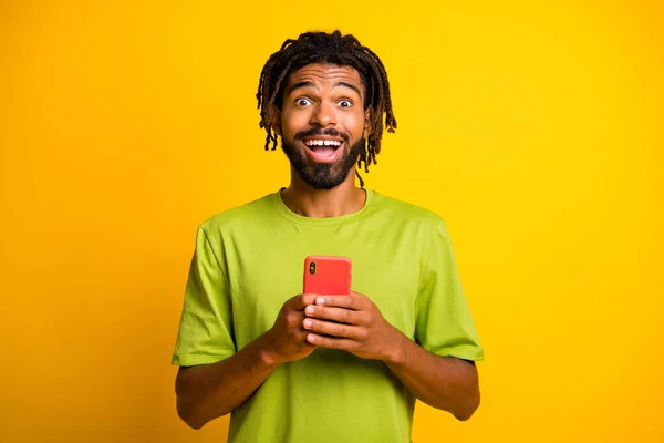 Foto van verbaasd donkere huid man houden smartphone onder de indruk dragen groene t-shirt geïsoleerd op glans gele kleur achtergrond — Stockfoto