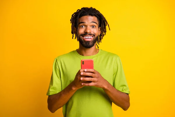 Foto de surpreendido cara pele escura segurar smartphone como conceito de reação desgaste camiseta verde isolado no fundo de cor amarela — Fotografia de Stock