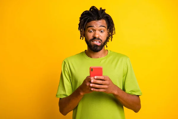 Endişeli koyu tenli adam sosyal medya bilgilerini okuyor cep telefonu ısırığı dişler sarı arka planda izole edilmiş tişört giyiyor — Stok fotoğraf
