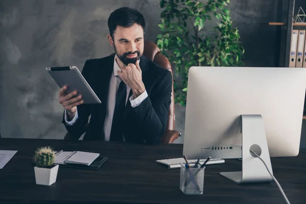 Foto de hombre exitoso sentarse escritorio mantenga tableta dedo barbilla mirada pc pantalla desgaste traje camisa corbata en la oficina moderna en interiores —  Fotos de Stock