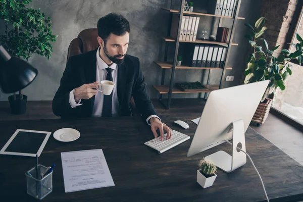 Foto av självsäker affärsman hålla mugg skriva tangentbord utseende skärm dator bära kostym skjorta slips i moderna kontor inomhus — Stockfoto