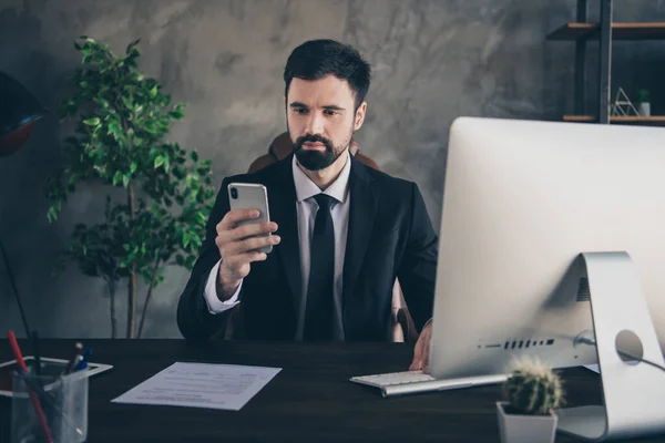 Фотографія успішного чоловіка сидить на столі комп'ютер тримає телефонний погляд екран читання смс одягу костюм сорочки краватка в сучасному офісі в приміщенні — стокове фото