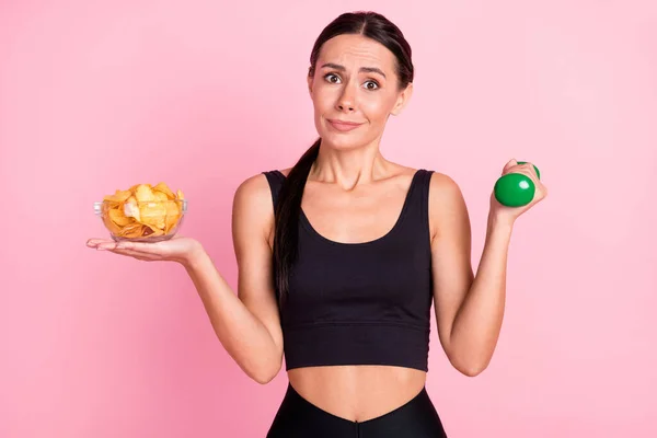 Foto de mujer joven insegura interrogada vestida con ropa deportiva sosteniendo patatas fritas mancuerna aislado color rosa fondo —  Fotos de Stock